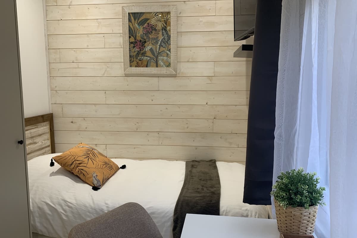 Un dormitorio con pared de madera, una cama con ropa blanca, una almohada y una mesita de noche, además de una pequeña planta en maceta.