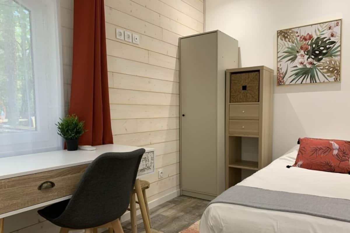 Un dormitorio con cama, escritorio, silla, armario y ventana con cortinas.