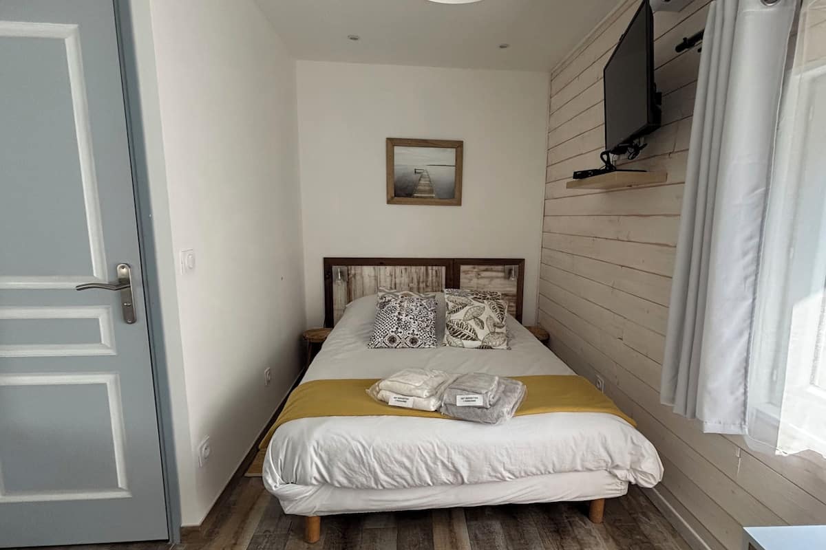 Un dormitorio con cama, almohadas, una mesita de noche, un televisor en una estantería, una puerta y una ventana.