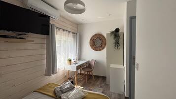 A compact room with a bed, a desk, a chair, a wall-mounted mirror, and a hanging plant.