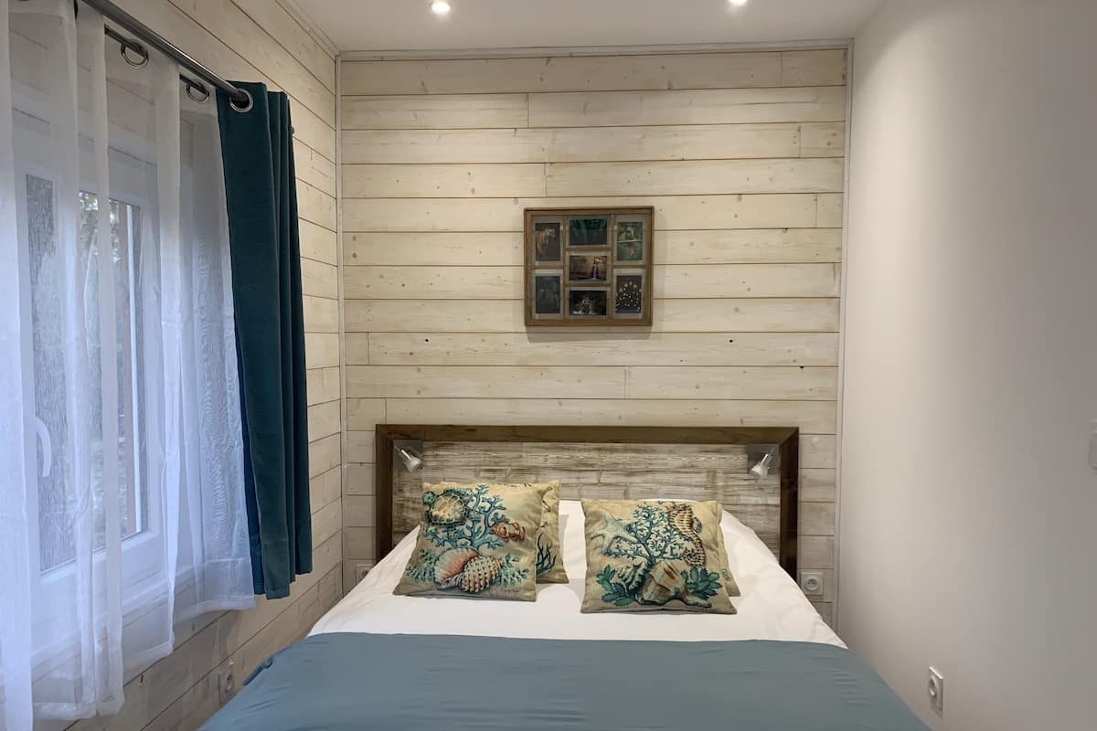 Un dormitorio con cabecera de madera, cama con dos almohadas decorativas, ventana con cortinas translúcidas y un pequeño cuadro en la pared.