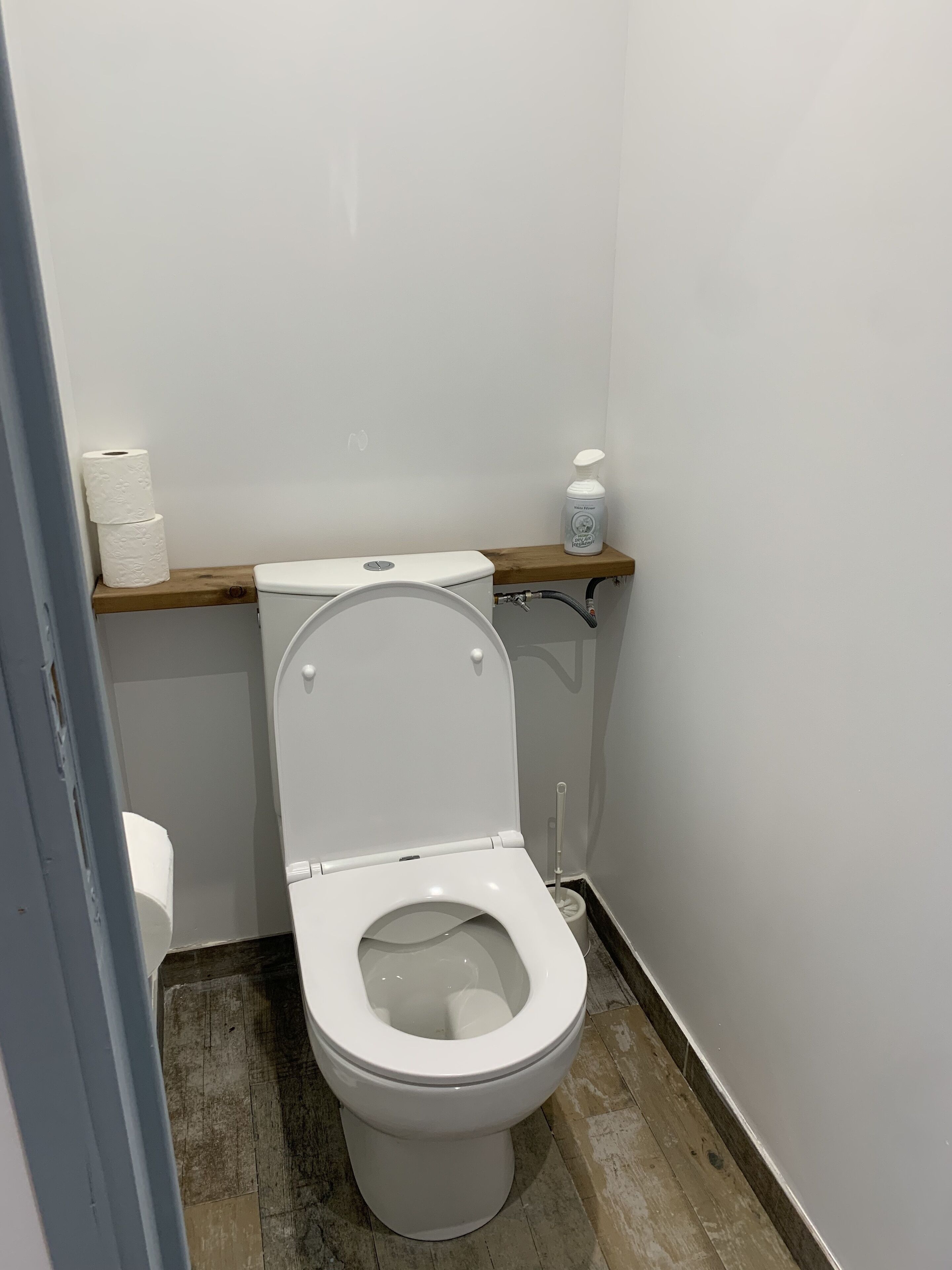 A small, clean bathroom with a white toilet, a wooden shelf with a bottle of hand soap, and a roll of toilet paper.