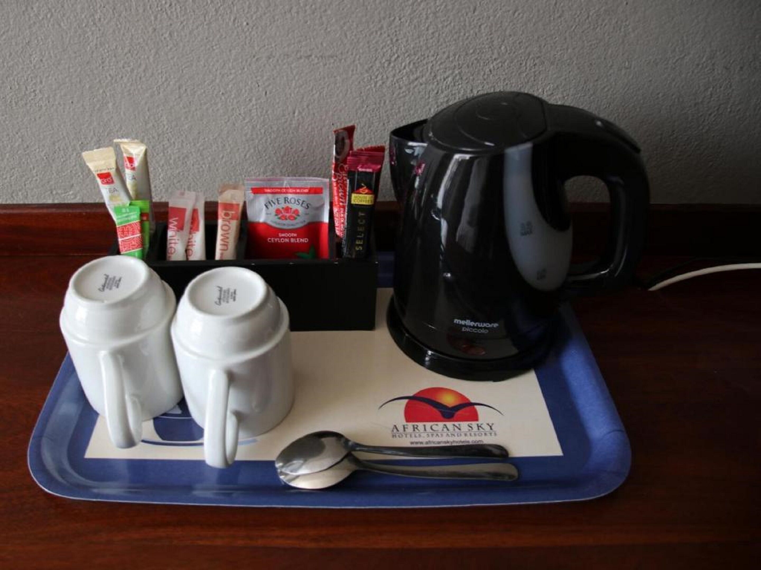 A tray with two white mugs, a spoon, a black coffee maker, and various coffee packets on a blue tray with a logo.