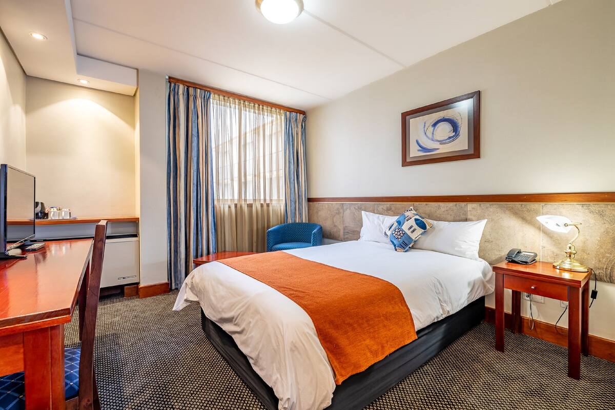 A hotel room with a bed, a desk, a chair, a nightstand, and a painting on the wall.