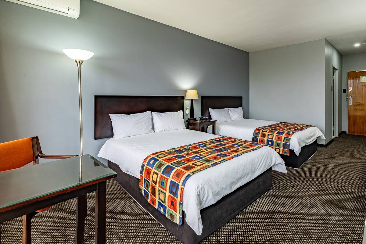 A hotel room with two beds, a desk, a chair, a lamp, and a TV.