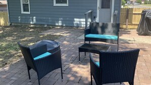 A patio with two wicker chairs and a small table, a house with a gray exterior, and a wooden fence in the background.