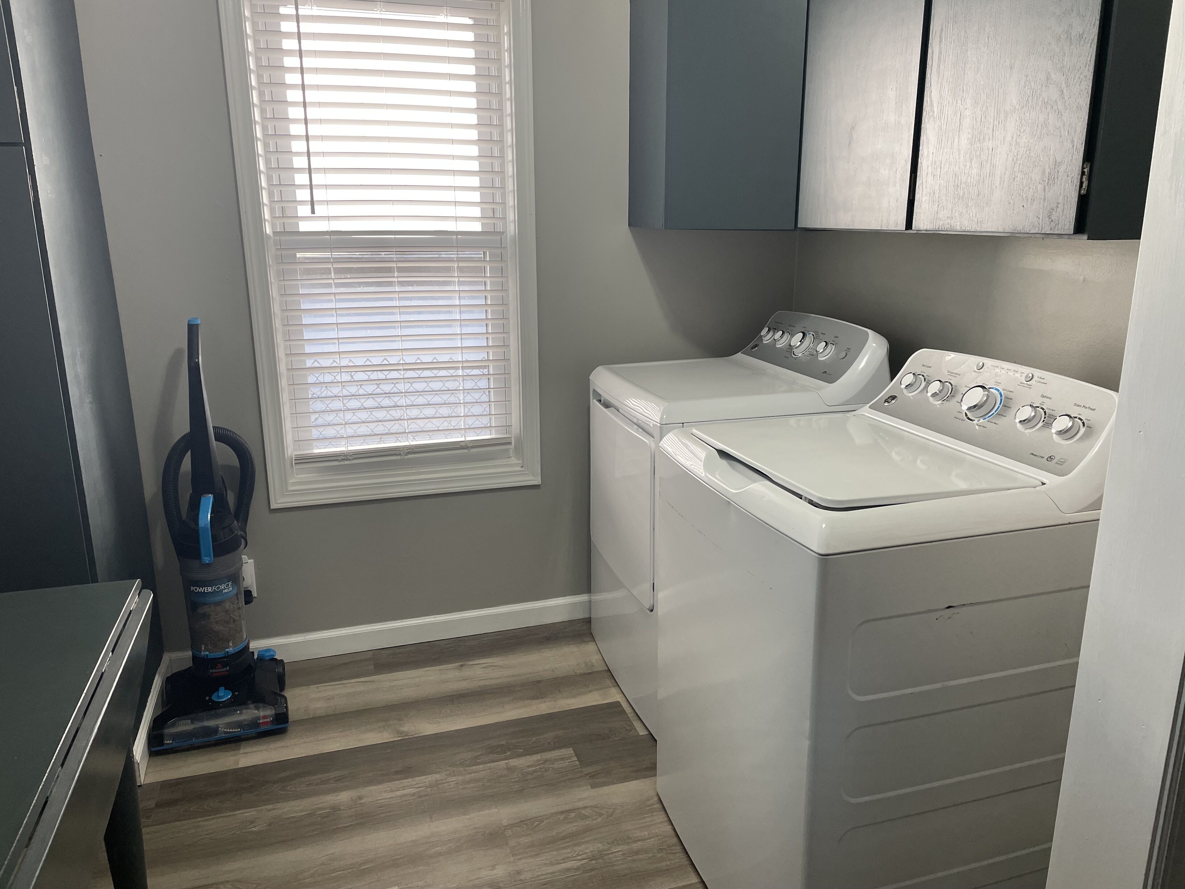 A laundry room with a front-loading washer and dryer, a vacuum cleaner, and a window with blinds.