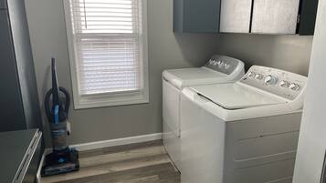 A laundry room with a front-loading washer and dryer, a vacuum cleaner, and a window with blinds.