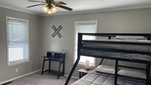 A room with a bunk bed, a desk, a ceiling fan, and two windows with blinds.
