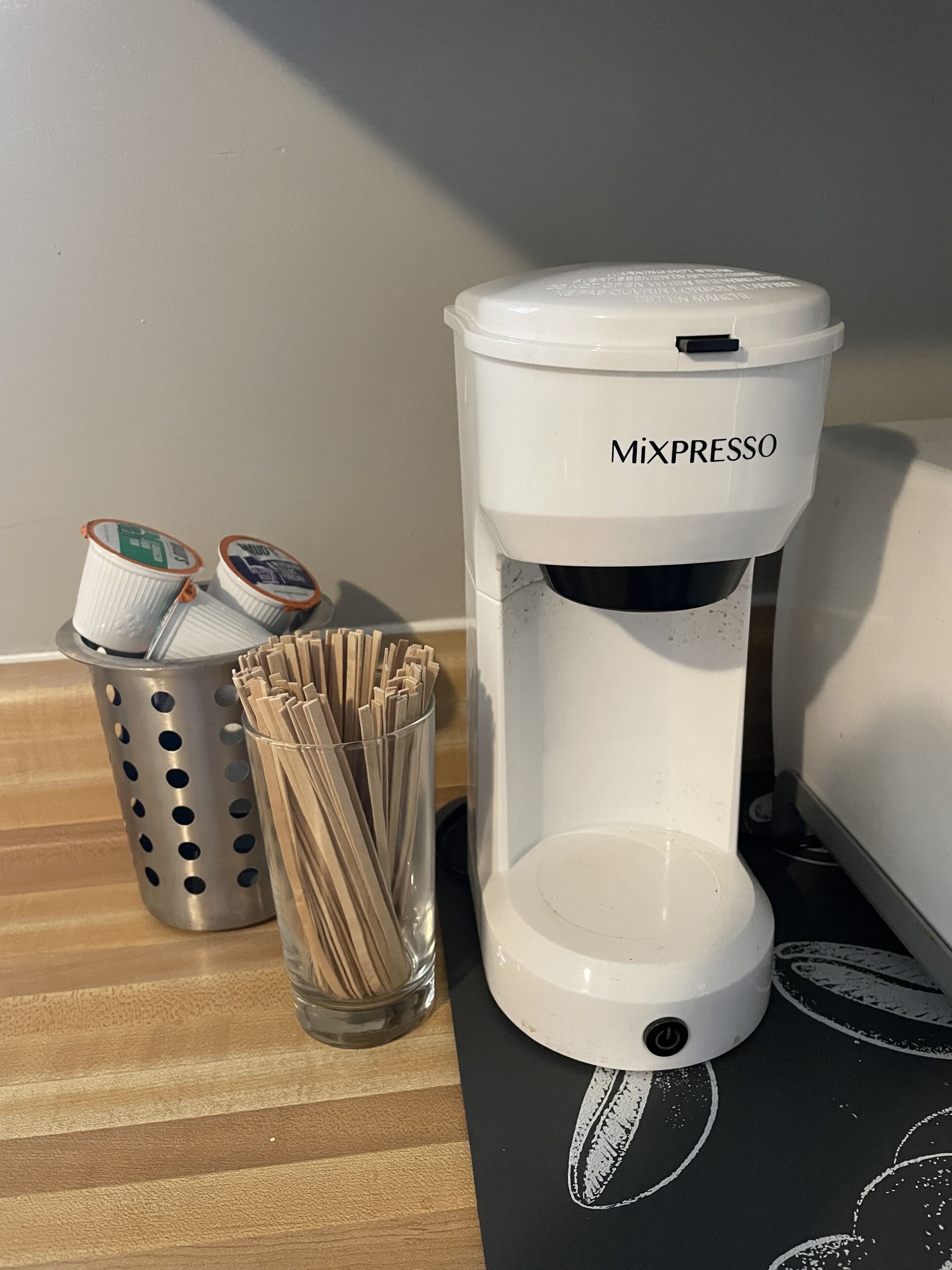 En kaffemaskin med etiketten ”MIXPRESSO” står på en bänkskiva, bredvid den finns en metallkorg med kaffekapslar och ett glas med sugrör.
