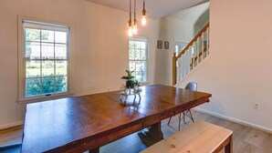 A dining area with a wooden table, bench, hanging pendant lights, and large windows with views of greenery.