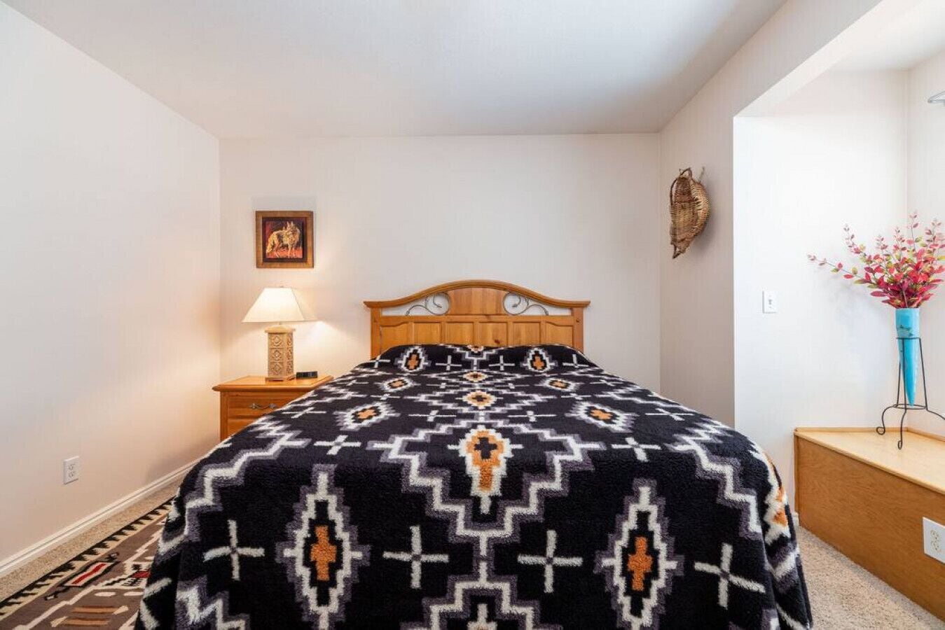 Un dormitorio con una cama de madera, una mesita de noche con una lámpara, una obra de arte en la pared, un adorno mural y un jarrón con flores.