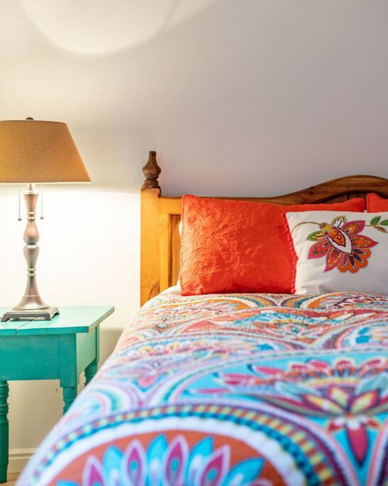 Un lit recouvert d’une couvre-lit aux motifs colorés, doté d’une tête de lit en bois et d’une table de chevet turquoise sur laquelle se trouve une lampe.