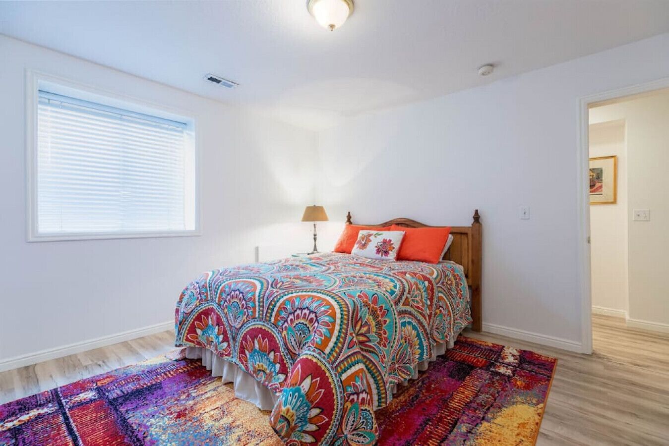 A bedroom with a colorful bedspread, a wooden headboard, a bedside lamp, and a patterned rug.
