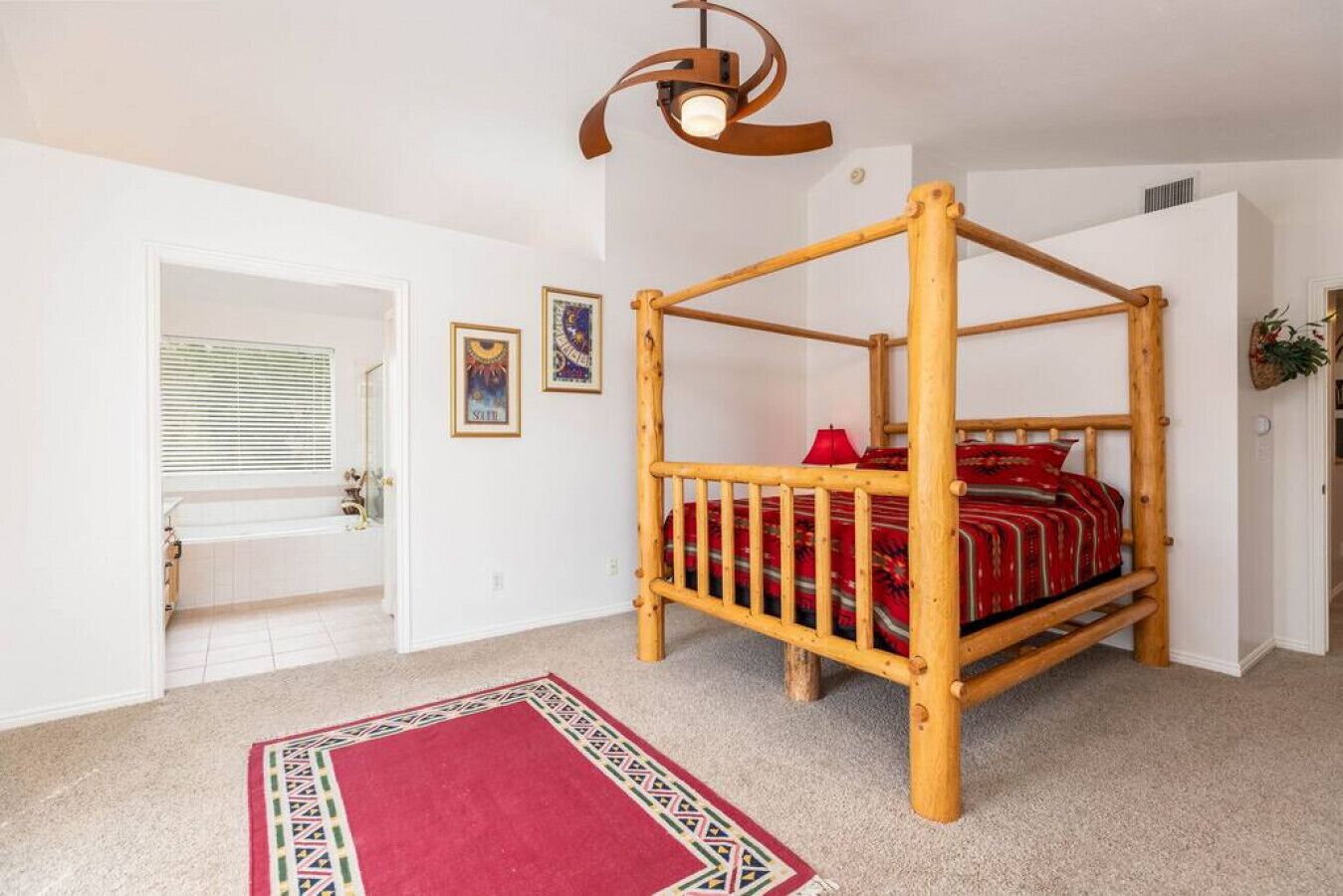 A room with a four-poster bed, a red and white patterned rug, a wooden ceiling fan, and a window with blinds.