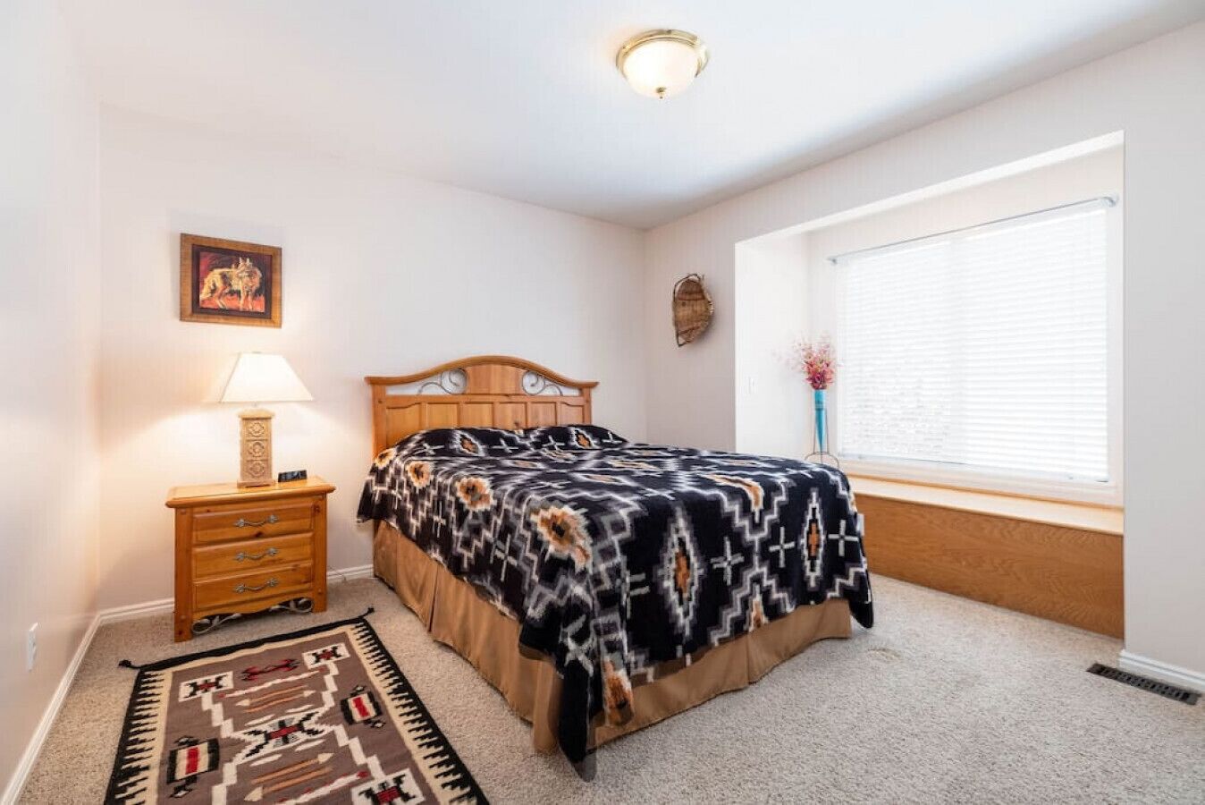 A bedroom with a bed, a nightstand, a lamp, a rug, a window with blinds, and a wall decoration.