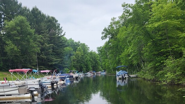 A boat docked at a marina with a row of other boats, surrounded by lush greenery. - Rustic and Eclectic open floor Plan Cabin with Canal view In Cranberry lake 😋.  (Harrison)