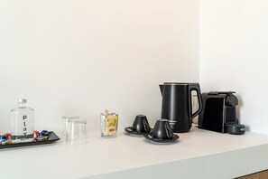 A hotel minibar setup with a bottle labeled 'PLAY,' a glass container with a yellow label, two black cups, a black coffee maker, and a black electric kettle.