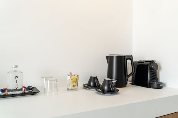 A hotel minibar setup with a bottle labeled 'PLAY,' a glass container with a yellow label, two black cups, a black coffee maker, and a black electric kettle.