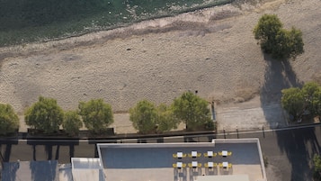 An aerial view of a coastal area with a sandy beach, a row of trees, and a building with outdoor seating.