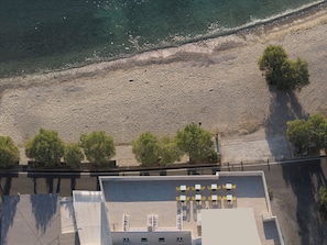 Vista aérea de una zona costera con una playa arenosa, una fila de árboles y un edificio con mesas al aire libre.