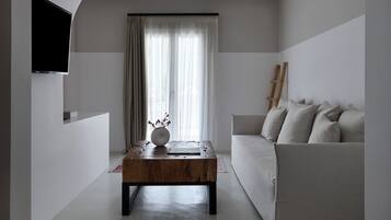 Una sala de estar minimalista con un sofá blanco, una mesa de centro de madera, un televisor de pantalla plana montado en la pared y un ventanal con cortinas de gasa.