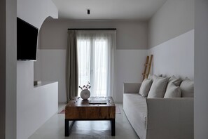 A minimalist living room with a white sofa, a wooden coffee table, a flat-screen TV mounted on the wall, and a large window with sheer curtains. - Play Paros, a member of Brown Hotels (Paros)