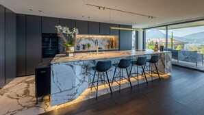 A modern kitchen with a marble island, dark cabinetry, and a large window offering a mountain view. - Villa Tomar (Talloires-Montmin)