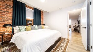 Een moderne slaapkamer met een accentmuur van baksteen, een groot bed, donkere gordijnen en uitzicht op de aangrenzende eetruimte.