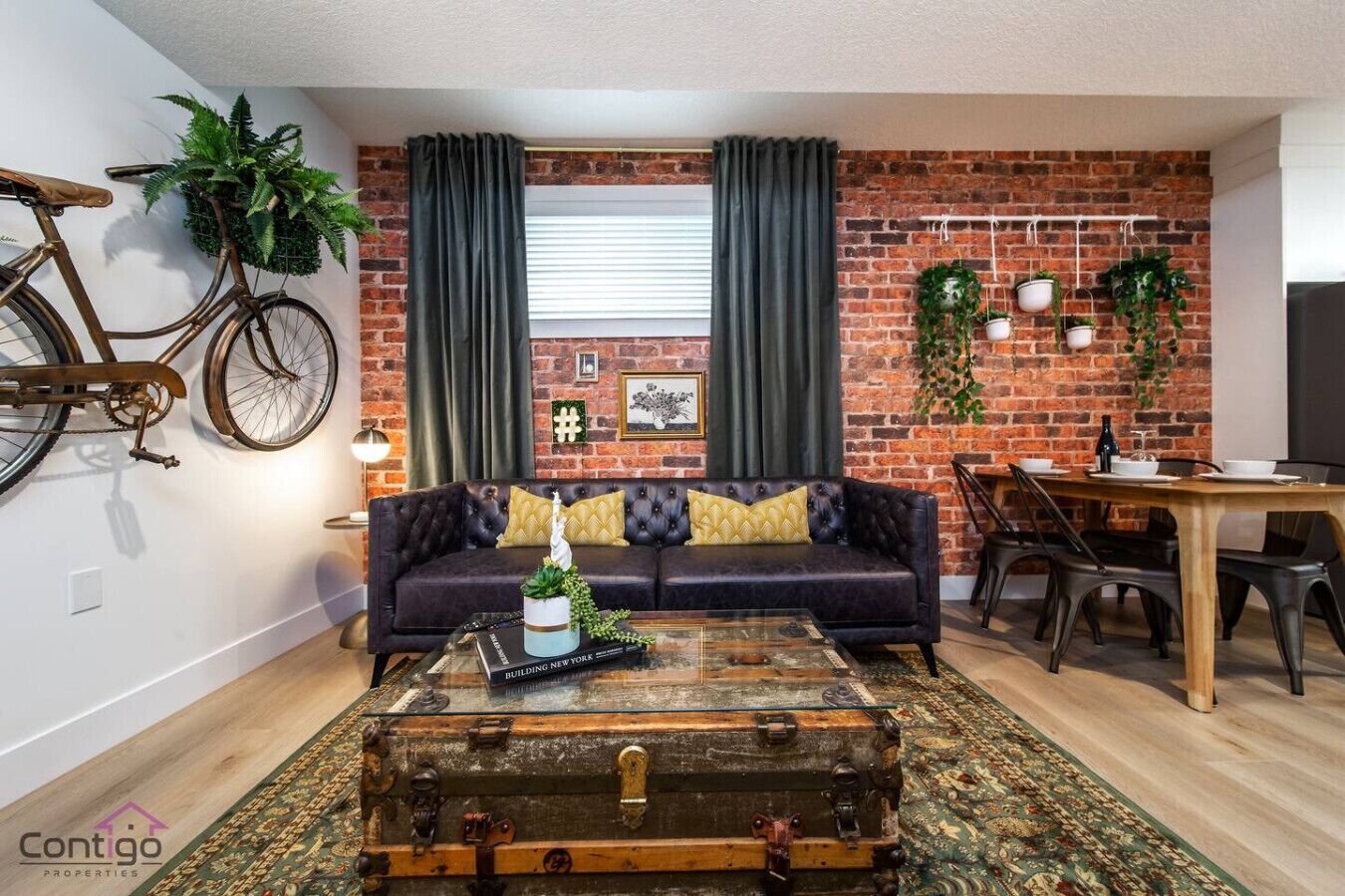 Uma sala de estar com sofá de couro, mesa de centro em formato de mala antiga, espaço de jantar com mesa e cadeiras de madeira, e uma parede de tijolos com plantas e uma bicicleta.