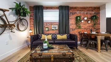 Uma sala de estar com sofá de couro, mesa de centro em formato de mala antiga, espaço de jantar com mesa e cadeiras de madeira, e uma parede de tijolos com plantas e uma bicicleta.