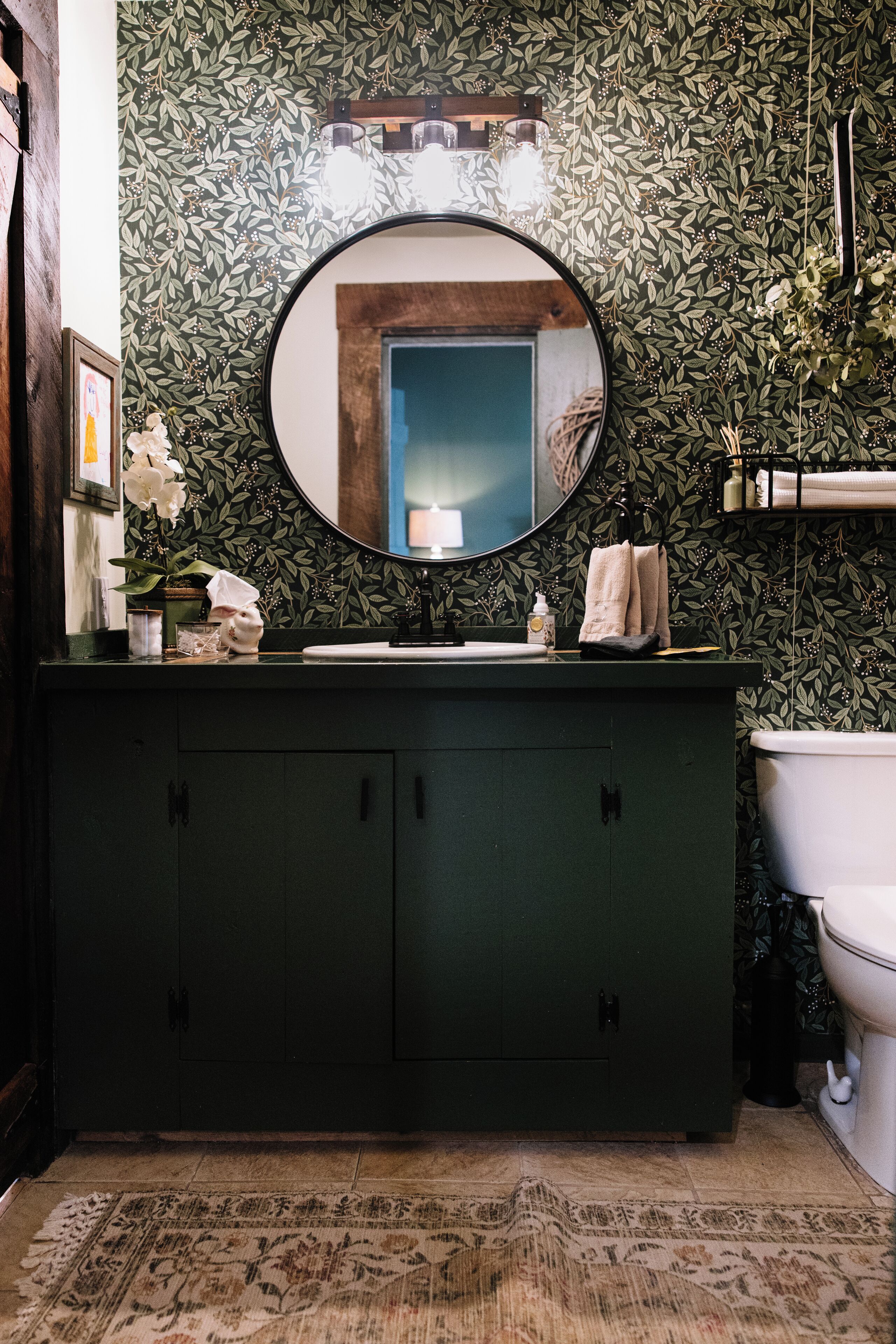 Une salle de bain avec un meuble-lavabo vert foncé, un miroir rond et un papier peint à motifs floraux.