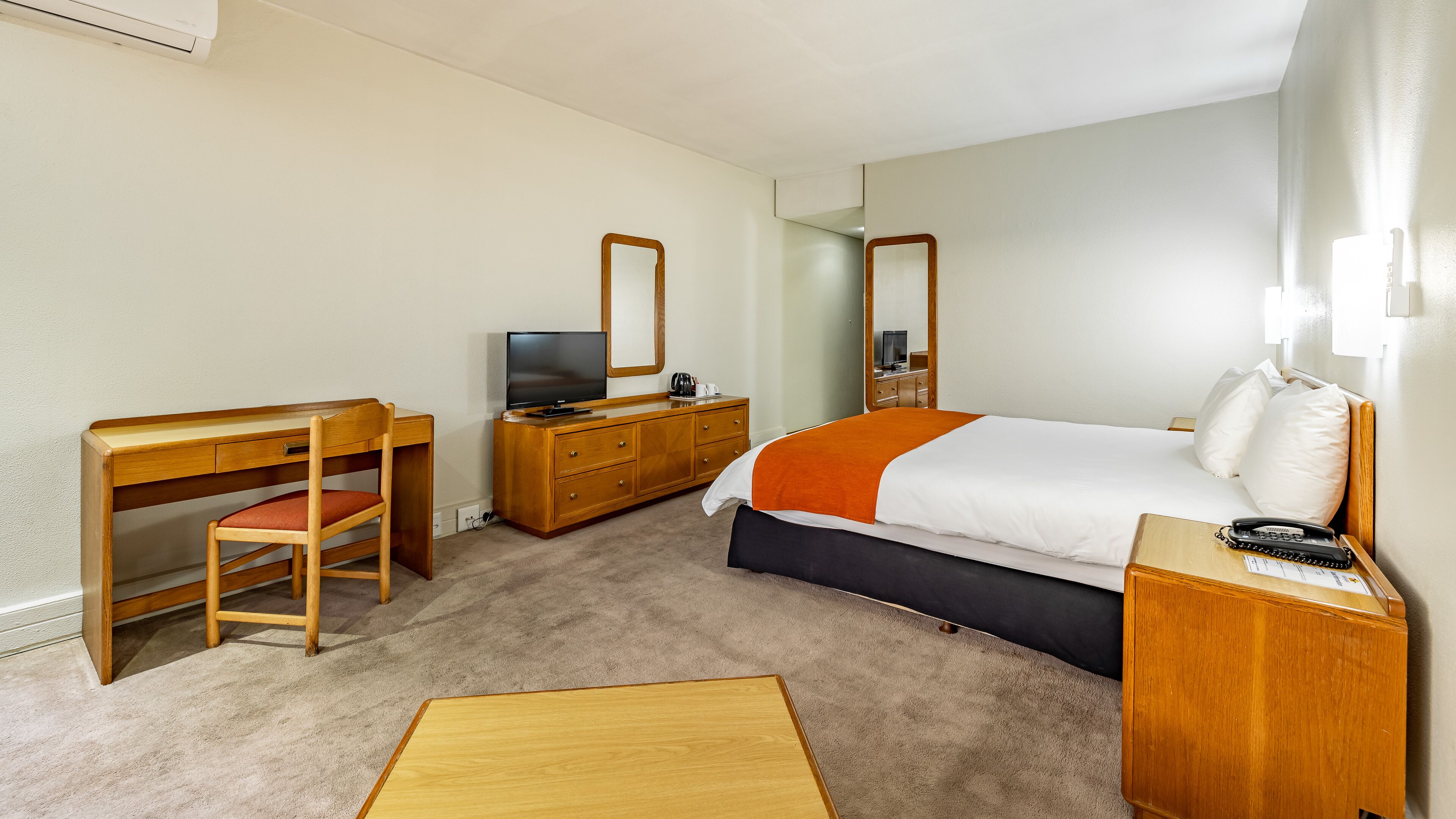 A hotel room with a bed, a desk with a chair, a dresser, a television, and a bedside table with a phone.