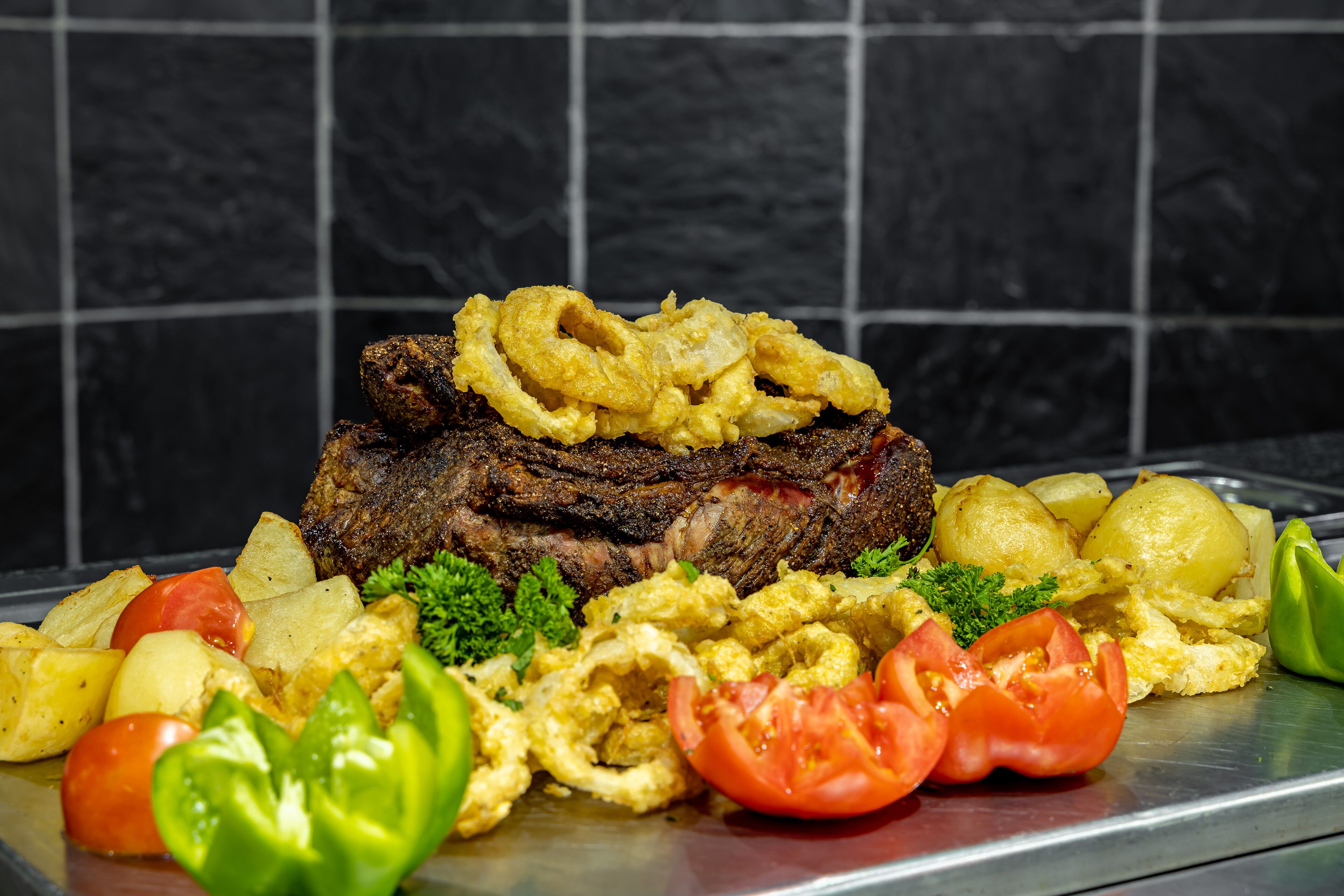 Ein Steak mit Zwiebelringen, Kartoffeln und Tomaten auf einer Metallplatte.