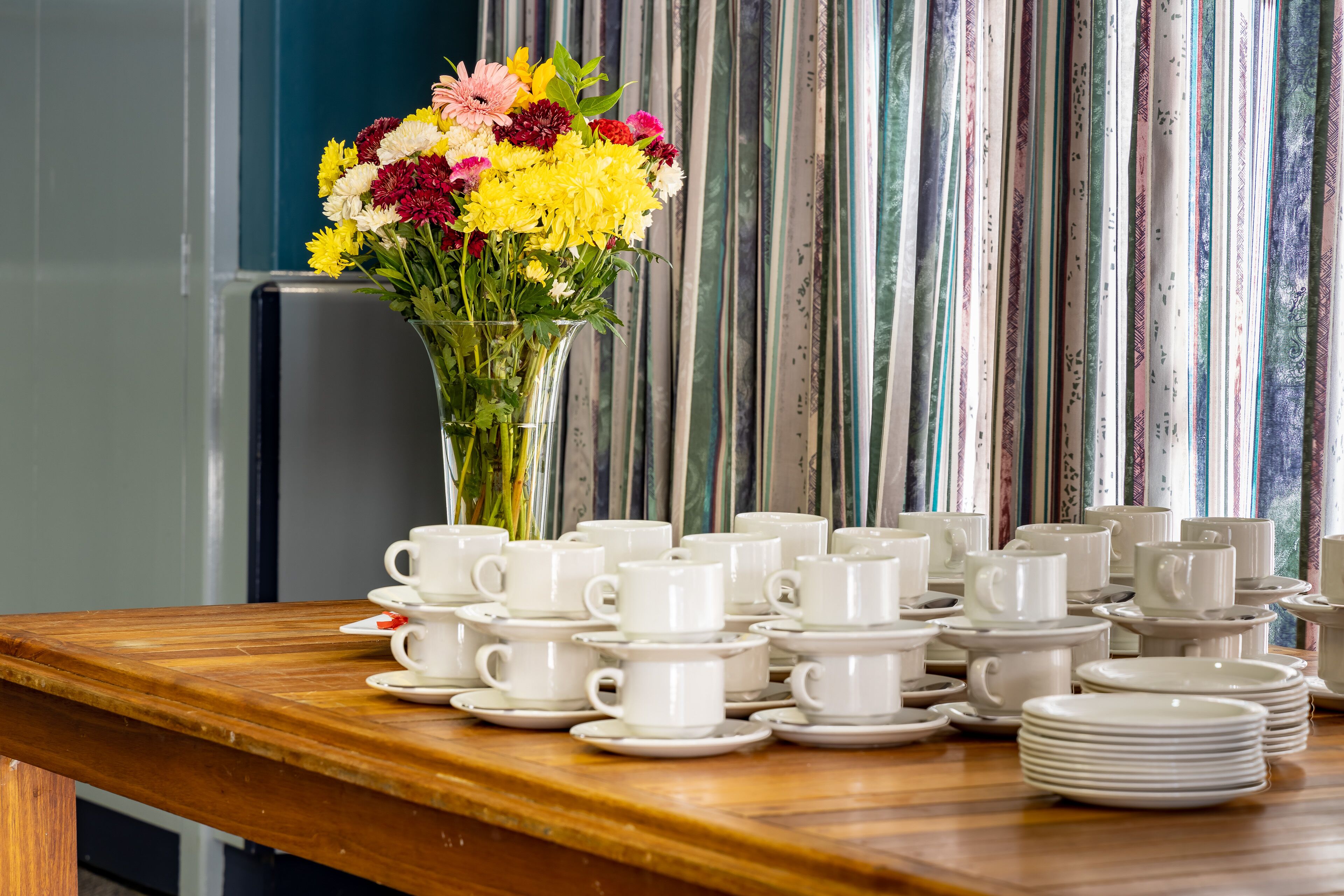 A table with white cups and saucers, a vase with yellow and pink flowers, and striped curtains in the background.