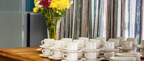 Una mesa con tazas y platillos blancos, un florero con flores amarillas y rosas, y cortinas a rayas al fondo.