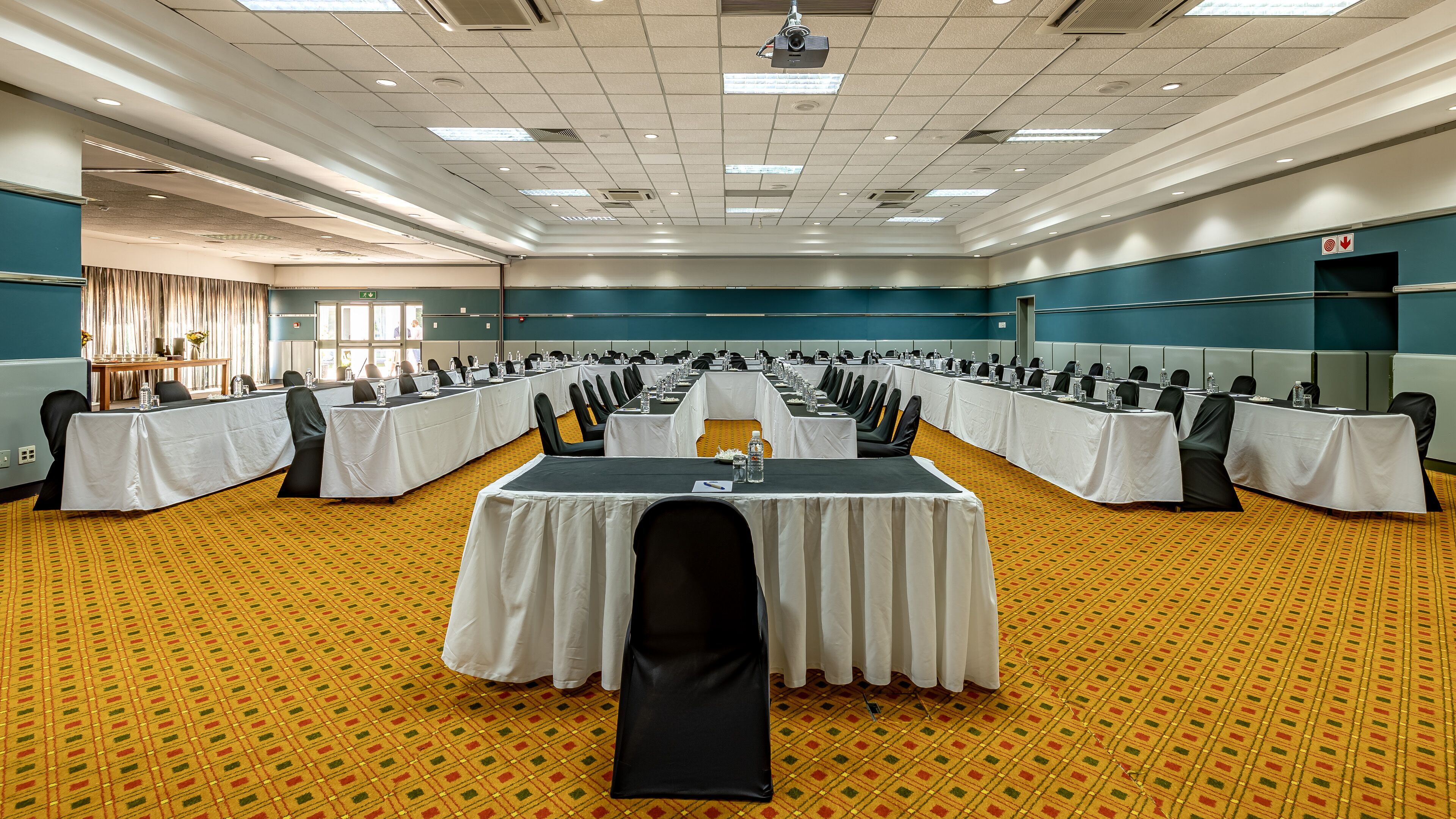 A large conference room with multiple tables and chairs, arranged in rows, with a carpeted floor and a high ceiling with recessed lighting.
