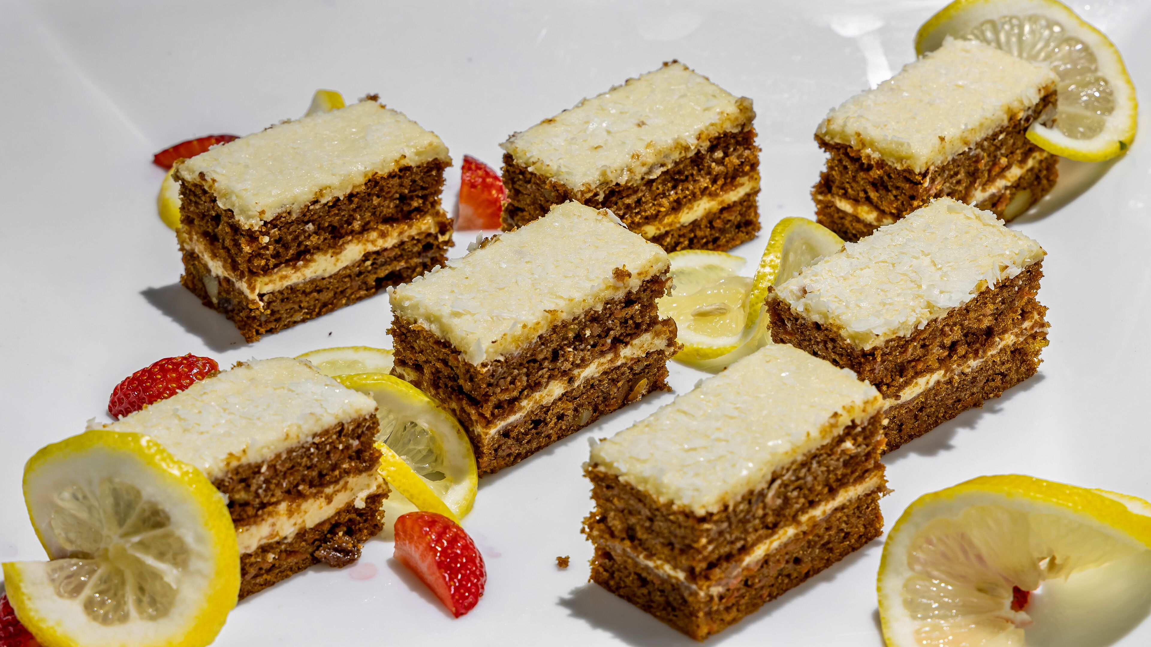Un plato blanco con varias porciones cuadradas de pastel en capas, decorado con rodajas de limón y fresas.