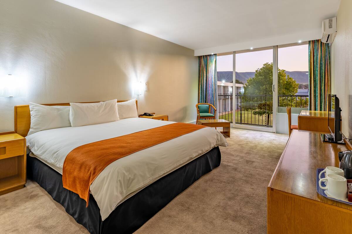 A hotel room with a large bed, a desk with a kettle, a chair, and a view of the outdoors.