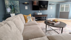 Un salón con un sofá color beige, una mesa de centro, un televisor de pantalla plana y un cuadro decorativo.