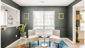 Una habitación con paredes verde oscuro, un ventanal con persianas, dos sillones, una mesita redonda, una planta en maceta y una alfombra colorida.