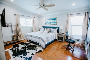 Une chambre à coucher comprenant un lit, un bureau, une chaise, un ventilateur de plafond et une fenêtre avec des rideaux.