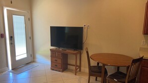 Una sala de estar con una televisión sobre un mueble de madera, una mesa de comedor con sillas, y una puerta con ventanal.