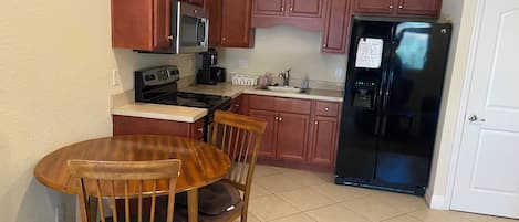 A compact kitchen with wooden cabinets, a black refrigerator, a microwave, a stove, and a sink. There is a wooden dining table with chairs.