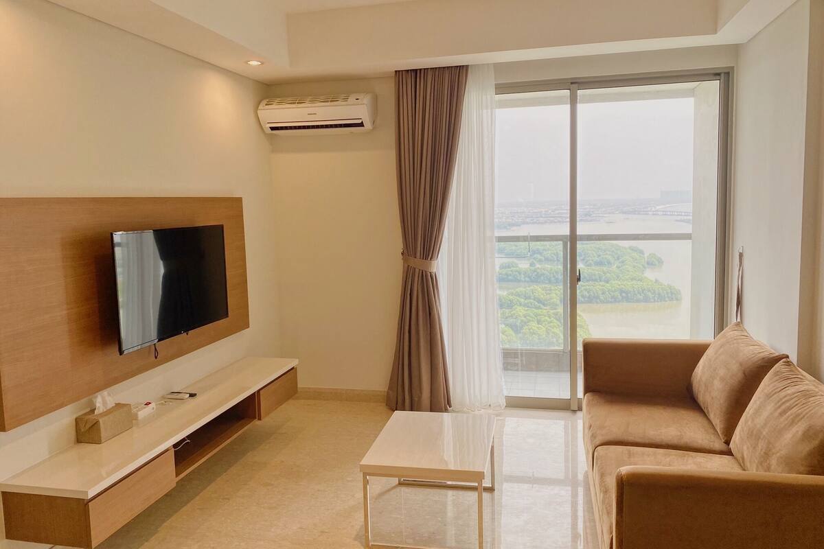 A modern living room with a flat-screen TV, a sofa, a coffee table, and a large window with a view of greenery and water.