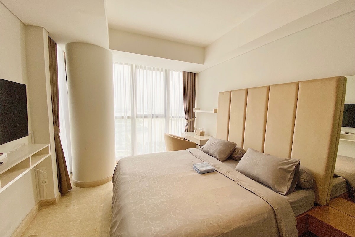 A hotel room with a large bed, a TV, a desk, and a window with curtains.