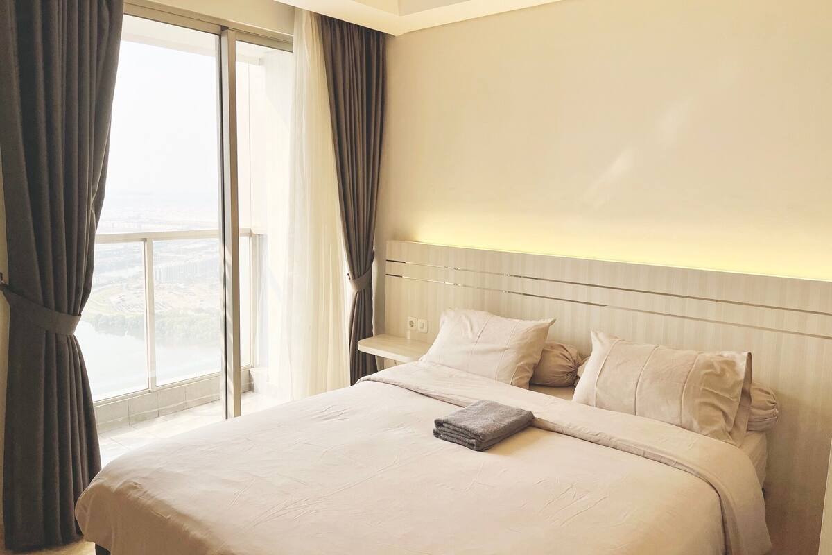 A hotel room with a large bed, a bedside table, and a view through a large window.