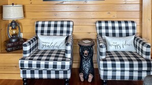 Interior acollidor d'una cabana amb dues butaques amb quadres, un quadre emmarcat d'un moll brumós, una taula petita amb una làmpada i una estàtua decorativa d'un gos.