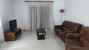 A living room with a brown leather sofa, a glass coffee table, a TV on a wooden stand, a lace curtain, and a red balloon. - Alojamento dos Redondos (Fernão Ferro)