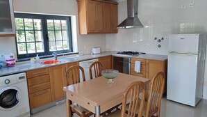 A kitchen with wooden cabinets, a sink, a dishwasher, a washing machine, a table with chairs, and a refrigerator. - Alojamento dos Redondos (Fernão Ferro)